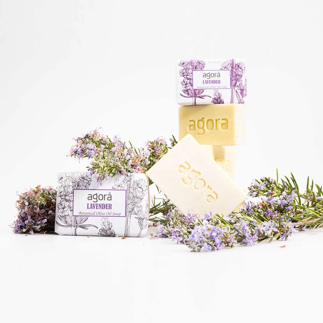 Stacked and wrapped bars of Soaperb Agorá Lavender Botanical Olive Oil Soap surrounded by blooming sprigs of lavender and rosemary on a white background.